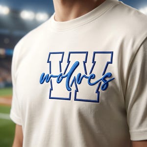 May include: A cream-colored short-sleeve t-shirt featuring a large, block-style letter W in blue outline. Overlaid across the center of the letter is the word wolves written in a blue, cursive, three-dimensional script font.