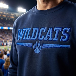 以下が含まれることがあります： 胸元に太い青色の文字でWILDCATSと刺繍されたネイビーブルーのクルーネックトレーナー。刺繍された青い肉球のグラフィックの上部に配置されており、肉球の両側には水平な線が伸びています。