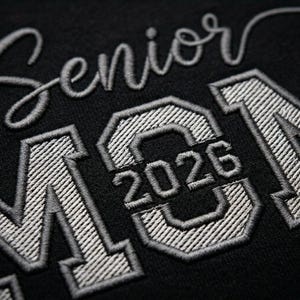 Extreme macro close-up of "Senior Mom 2026" embroidery on a black cotton t-shirt. This high-detail shot captures the specific stitch direction, fabric grain, and luxury thread surface, providing a clear view of the high-quality digitizing work.