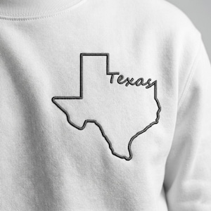 May include: White sweatshirt featuring an embroidered outline of the state of Texas in dark gray. The word "Texas" is embroidered in a cursive font within the state's borders. The sweatshirt is made of a soft, comfortable material.