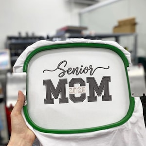 Puede incluir: Tela blanca con las palabras "Senior MOM 2026" bordadas en gris y negro. La tela se sujeta en un bastidor de bordado verde. El texto est&aacute; en una fuente de escritura y letras de bloque.