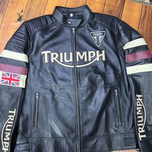 May include: A black leather motorcycle jacket featuring a stand-up collar with a snap button, a full-length front zipper, and two zippered waist pockets. The chest displays the word Triumph in white, with a Triumph logo patch above it.