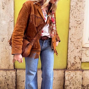 May include: A brown suede jacket with fringe detailing, worn with a matching crossbody bag. The jacket is over a white floral shirt and blue jeans. A patterned scarf and belt complete the look, creating a bohemian style.