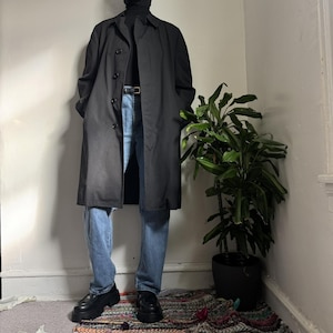 May include: A black trench coat with a button-up front, worn over a black roll neck and blue jeans. The outfit includes black platform shoes and a black belt. A potted plant is in the background.