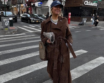 May include: A person wearing a brown suede trench coat, blue jeans, and a navy baseball cap, crossing a city street. They carry a newspaper and a patterned handbag. The background features buildings, cars, and a restaurant named "Chinato".