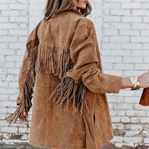 May include: A brown suede jacket with fringing on the back and sleeves. The jacket features a classic design with a collared neckline and pockets. The fringing adds a bohemian flair. The jacket is a medium length, perfect for layering.
