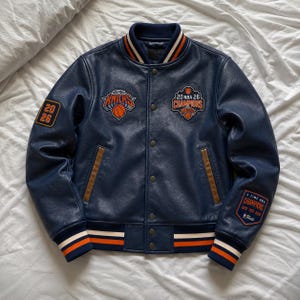 Pode incluir: Uma jaqueta universitária azul-marinho com o logotipo dos New York Knicks, um emblema 20 NBA 26 Champions e um emblema 3 Time NBA Champions 1970 1973 2026 Finals. A jaqueta tem fecho de botões de pressão e gola, punhos e bainha canelados com listras laranja e brancas.