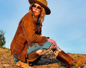 May include: A person wearing a brown suede fringed jacket, a tan cowboy hat, and brown sunglasses. The person is also wearing a denim dress and brown cowboy boots. The person is squatting on a rocky surface under a blue sky.