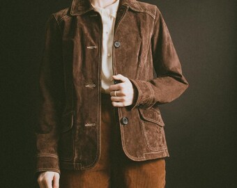 May include: A brown suede jacket with a button-up front, two front pockets, and a collared neckline. The jacket is paired with matching brown corduroy pants. The outfit is a vintage style.