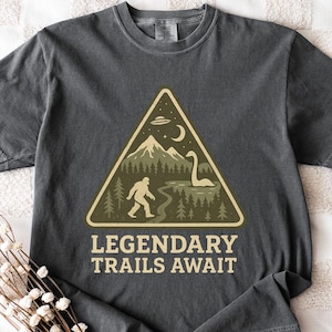 Op de afbeelding: Donkergrijs t-shirt met een beige driehoekige afbeelding met een bergketen, Bigfoot en een meer met een wezen. De tekst "LEGENDARY TRAILS AWAIT" staat eronder. Het shirt ligt plat op een wit patroon.