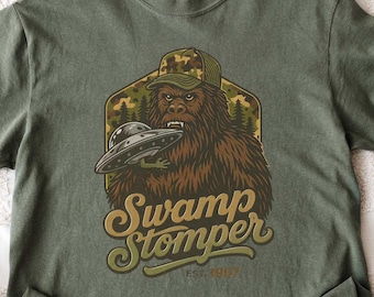 May include: Olive green t-shirt featuring a Bigfoot graphic wearing a camouflage hat and holding a UFO. The text "Swamp Stomper" is printed in a vintage style. The shirt is folded on a white checkered surface.