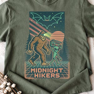 May include: Olive green t-shirt featuring a graphic of Bigfoot and an alien walking together, with a retro sunset and bat design. The text "MIDNIGHT HIKERS" is displayed below the graphic.