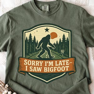 Op de afbeelding: Olijfgroen t-shirt met een Bigfoot-afbeelding in een schildvormig ontwerp. De afbeelding toont een Bigfoot-silhouet, bomen en de tekst "Sorry I'm late - I saw Bigfoot". Het shirt is opgevouwen op een wit patroon.