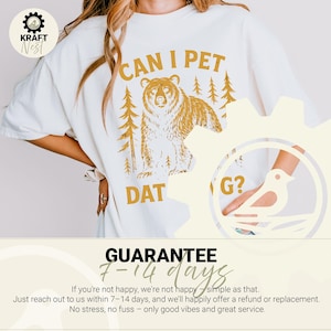 May include: Cream-colored t-shirt with a gold bear graphic and the text "CAN I PET DAT G?". The design includes trees, and the shirt has a relaxed fit. A guarantee is also printed on the bottom of the shirt.