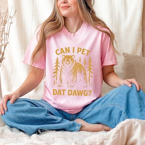 May include: Pink t-shirt with the text "CAN I PET DAT DAWG?" in gold lettering. The graphic features a bear and two trees. The shirt is being worn by a person wearing blue jeans.