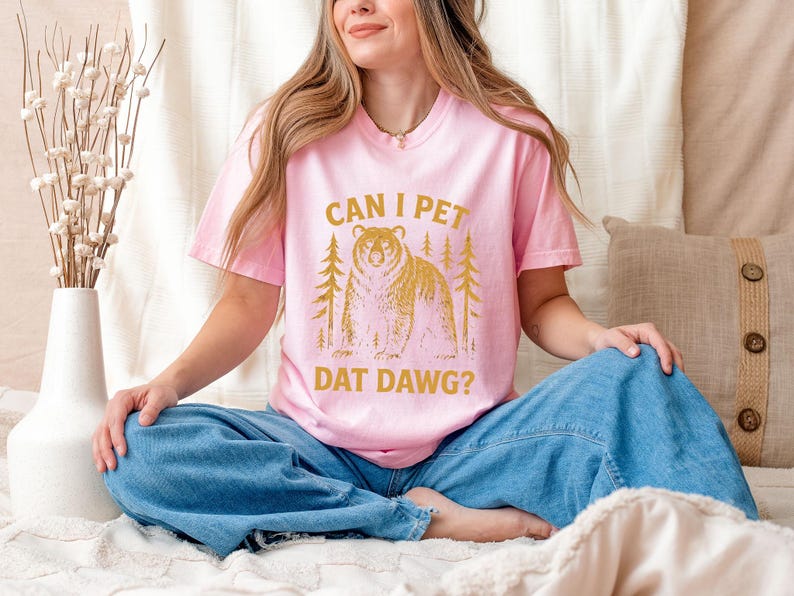 May include: Pink t-shirt with the text "CAN I PET DAT DAWG?" in gold lettering. The graphic features a bear and two trees. The shirt is being worn by a person wearing blue jeans.