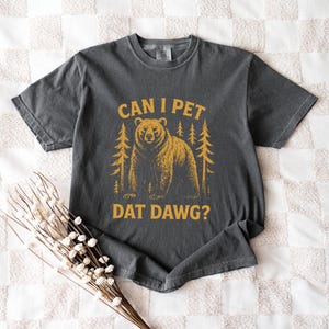 May include: Dark gray t-shirt with a distressed graphic featuring a bear and pine trees. The text "CAN I PET DAT DAWG?" is printed in a gold color. The shirt is laid flat on a white quilted surface.