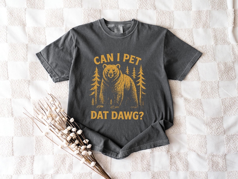May include: Dark gray t-shirt with a distressed graphic featuring a bear and pine trees. The text "CAN I PET DAT DAWG?" is printed in a gold color. The shirt is laid flat on a white quilted surface.