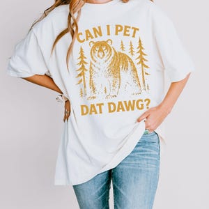 May include: Cream-colored t-shirt with a bear graphic and the text "CAN I PET DAT DAWG?". The bear is brown and is surrounded by pine trees. The shirt is a relaxed fit and is paired with blue jeans.