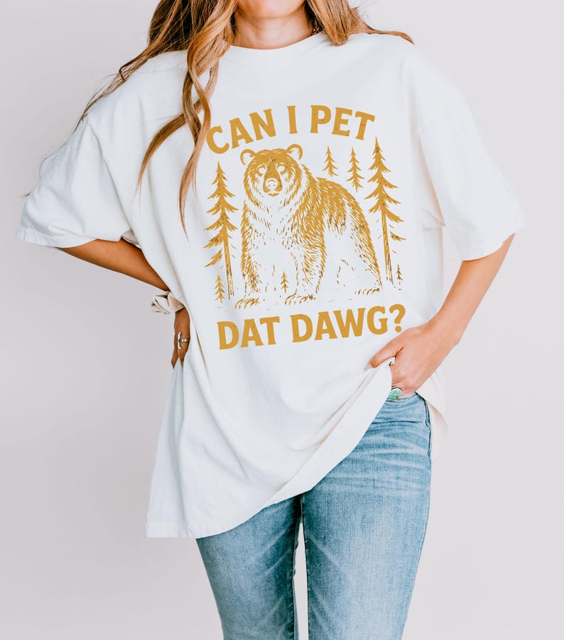 May include: Cream-colored t-shirt with a bear graphic and the text "CAN I PET DAT DAWG?". The bear is brown and is surrounded by pine trees. The shirt is a relaxed fit and is paired with blue jeans.