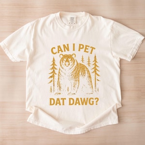 May include: Cream-colored t-shirt with a gold graphic of a bear and pine trees. The text "CAN I PET DAT DAWG?" is printed in gold. The shirt is made of a soft material and has short sleeves.