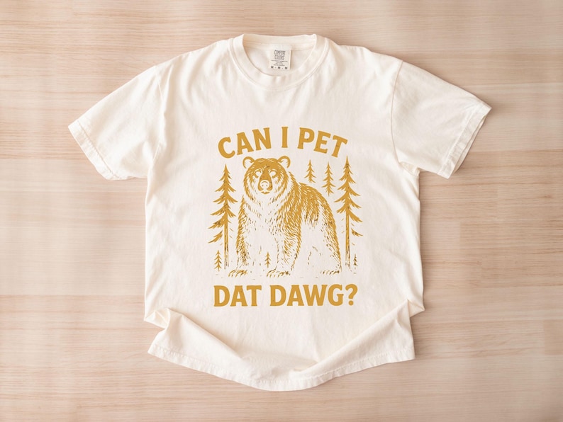 May include: Cream-colored t-shirt with a gold graphic of a bear and pine trees. The text "CAN I PET DAT DAWG?" is printed in gold. The shirt is made of a soft material and has short sleeves.