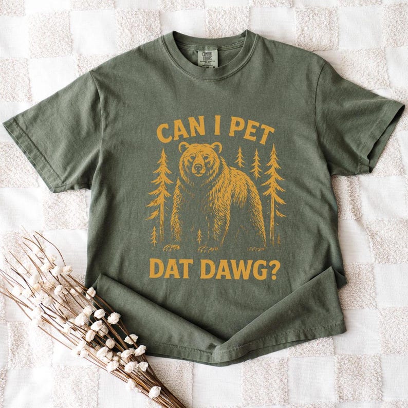 May include: Olive green t-shirt with the text "CAN I PET DAT DAWG?" in yellow. A yellow bear and pine trees are printed below the text. The shirt is folded on a white and cream checkered surface.