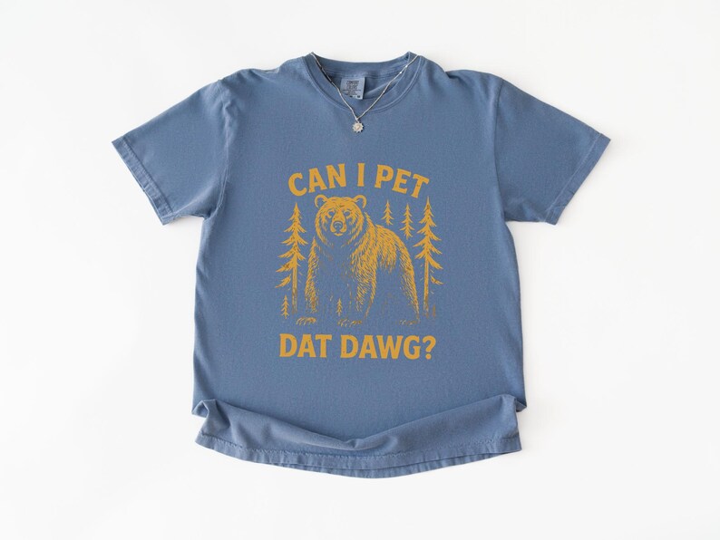 May include: A blue t-shirt with the text "CAN I PET DAT DAWG?" in yellow. The graphic features a bear and two trees. The shirt has short sleeves and a crew neck.