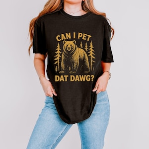May include: Black t-shirt with a gold graphic of a bear and pine trees. The text "CAN I PET DAT DAWG?" is above and below the bear illustration. The shirt is worn by a person wearing blue jeans.