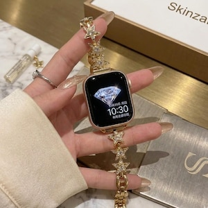 May include: Gold-toned smartwatch with a diamond-shaped display. The watch band features a series of star-shaped embellishments with clear crystals. The watch is held in a hand with long, manicured nails. The brand name "Skinzal" is visible.