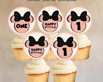 May include: Cupcakes topped with round toppers featuring a black bow, mouse ears, and the words "ONE", "HAPPY BIRTHDAY", and the number "1". The toppers are pink and cream colored. The cupcakes have yellow cake and white frosting.
