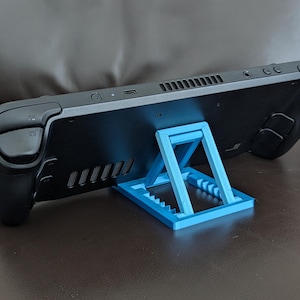 Op de afbeelding: Een zwarte handheld gameconsole wordt ondersteund door een lichtblauwe standaard. De standaard is gemaakt van plastic en heeft een geometrisch ontwerp. De console rust op een bruin oppervlak.