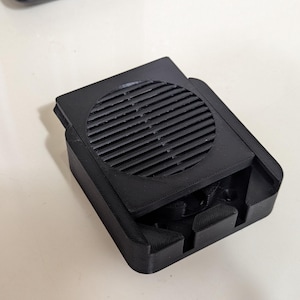 Può includere: Un dispositivo nero e rettangolare con una presa d'aria circolare a lamelle sulla parte superiore. Il dispositivo ha un'area incassata e sembra essere in plastica. Il design è funzionale e moderno.