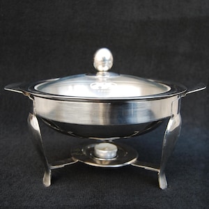 Puede incluir: Un chafing dish plateado con tapa de cristal transparente y un pequeño asa. El plato tiene un cuerpo redondeado y está sostenido por tres patas curvas. Una pequeña vela se encuentra debajo. La tapa está marcada con "22cm".