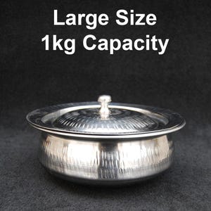 Puede incluir: Un bol de servir de acero inoxidable plateado con tapa. El bol tiene un acabado martillado y una capacidad de 1 kg. El texto "Large Size 1kg Capacity" está impreso sobre el bol.