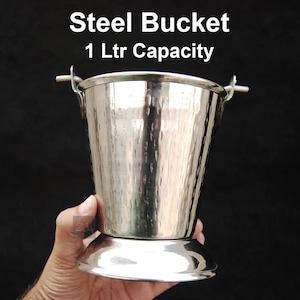 Puede incluir: Un cubo de acero inoxidable con textura martillada, con una capacidad de 1 litro. El cubo tiene forma acampanada, base redondeada y asas pequeñas. El texto "Steel Bucket 1 Ltr Capacity" se muestra en la parte superior.