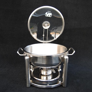 May include: A stainless steel chafing dish with a clear glass lid. The lid is slightly ajar, revealing the interior of the dish. The chafing dish has two handles and sits on a stand with a heating element.
