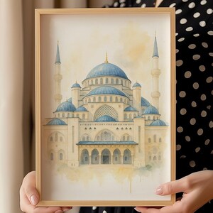 May include: A framed watercolor print of a mosque with blue domes and minarets. The building is painted in shades of beige and blue, with intricate architectural details. The frame is made of light-colored wood.