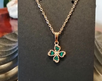May include: Gold-toned necklace with a four-leaf clover pendant. The clover features green heart-shaped accents and is outlined with small, clear crystals. The chain and pendant are displayed against a black background.