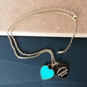 May include: Gold-colored necklace with two heart-shaped pendants. One pendant is a bright turquoise color, and the other is gold with the words "PLEASE RETURN TO FOREVER LOVE NEW YORK" engraved on it. The necklace is on a dark blue surface.