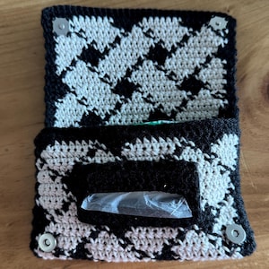 Pode incluir: Uma carteira de crochê feita à mão com um padrão de pata de galo em preto e branco. A carteira tem um fecho de aba com dois fechos de pressão prateados. O interior é visível e um pequeno item está dentro de uma embalagem plástica transparente.