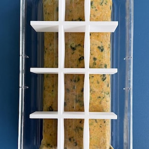 以下が含まれることがあります： 白い格子状のインサートが付いた透明なプラスチック製の食品保存容器で、いくつかの密封された食品の袋が入っています。食べ物は野菜のピューレのようです。容器は青い表面に置かれています。