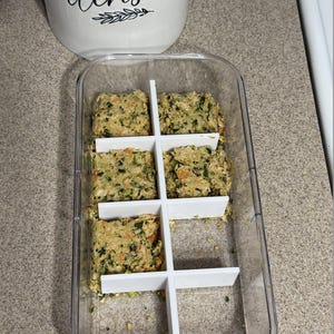 May include: A clear plastic container with a white divider holding six portions of a green and tan food mixture. A white utensil holder with the word "utensils" is in the background.