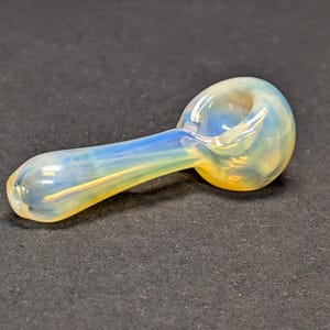 Op de afbeelding: Een handgeblazen glazen pijp met een afgeronde kom en een slanke steel. De pijp heeft een mix van kleuren, waaronder geel, blauw en helder, waardoor een opalescent effect ontstaat. Het glas is glad en lijkt goed gemaakt.