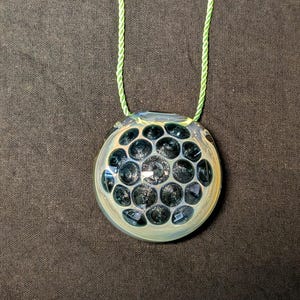May include: A round glass pendant with a pattern of dark blue bubbles, suspended by a light green cord. The pendant has a light blue and yellow rim, and the background is a dark gray fabric.