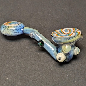 Peut inclure: Une pipe en verre soufflé à la main, aux tons bleus et blancs. Le foyer présente un tourbillon de couleurs arc-en-ciel. La pipe a une longue tige avec un petit embout buccal rond. Le design comprend de petits accents décoratifs en verre.