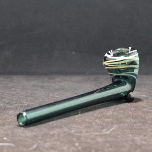 Peut inclure: Une pipe en verre vert avec des motifs en spirale blancs. La pipe a une longue tige incurvée et un bol en haut.