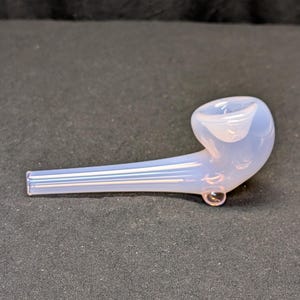 Peut inclure: Une pipe en verre bleu clair avec un foyer arrondi et une longue tige droite. La pipe a une finition claire et lisse avec des stries blanches. Une petite bulle de verre transparente est au fond du foyer.