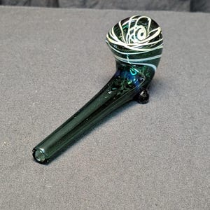 Peut inclure: Une pipe en verre vert foncé avec un foyer arrondi orné de tourbillons blancs et verts. La tige est longue et droite, avec un petit pied à la base. La pipe est conçue pour fumer.