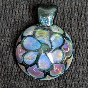 Może przedstawiać: Okrągła, iryzująca szklana zawieszka z pętelką u góry. Zawieszka ma kwiatowy wzór w odcieniach niebieskiego, fioletowego i zielonego, z błyszczącym, teksturowanym wykończeniem. Tło to ciemna, teksturowana tkanina.